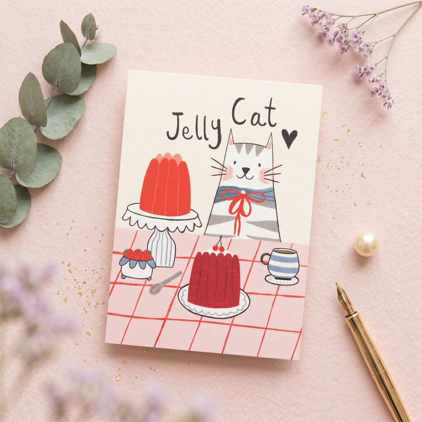 A cute illustrated greeting card titled 'Jelly Cat' lies on a pink surface with decorative eucalyptus leaves, small purple flowers, a pearl-like bead, and a gold pen. The card shows a gray-and-white cat wearing a red bow around its neck, standing beside red jelly cakes and a striped cup on a pink checkered tablecloth.