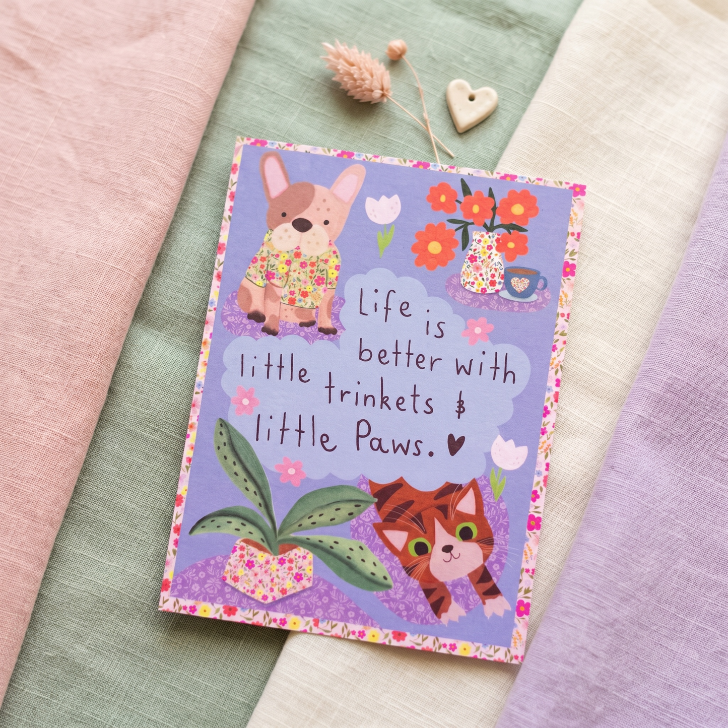 A square greeting card featuring cute illustrations of a French bulldog and a tabby cat with the text 'Life is better with little trinkets & little Paws. ♥' on a lavender background, styled on a soft fabric surface with pink, sage green, cream, and lavender cloth layers.