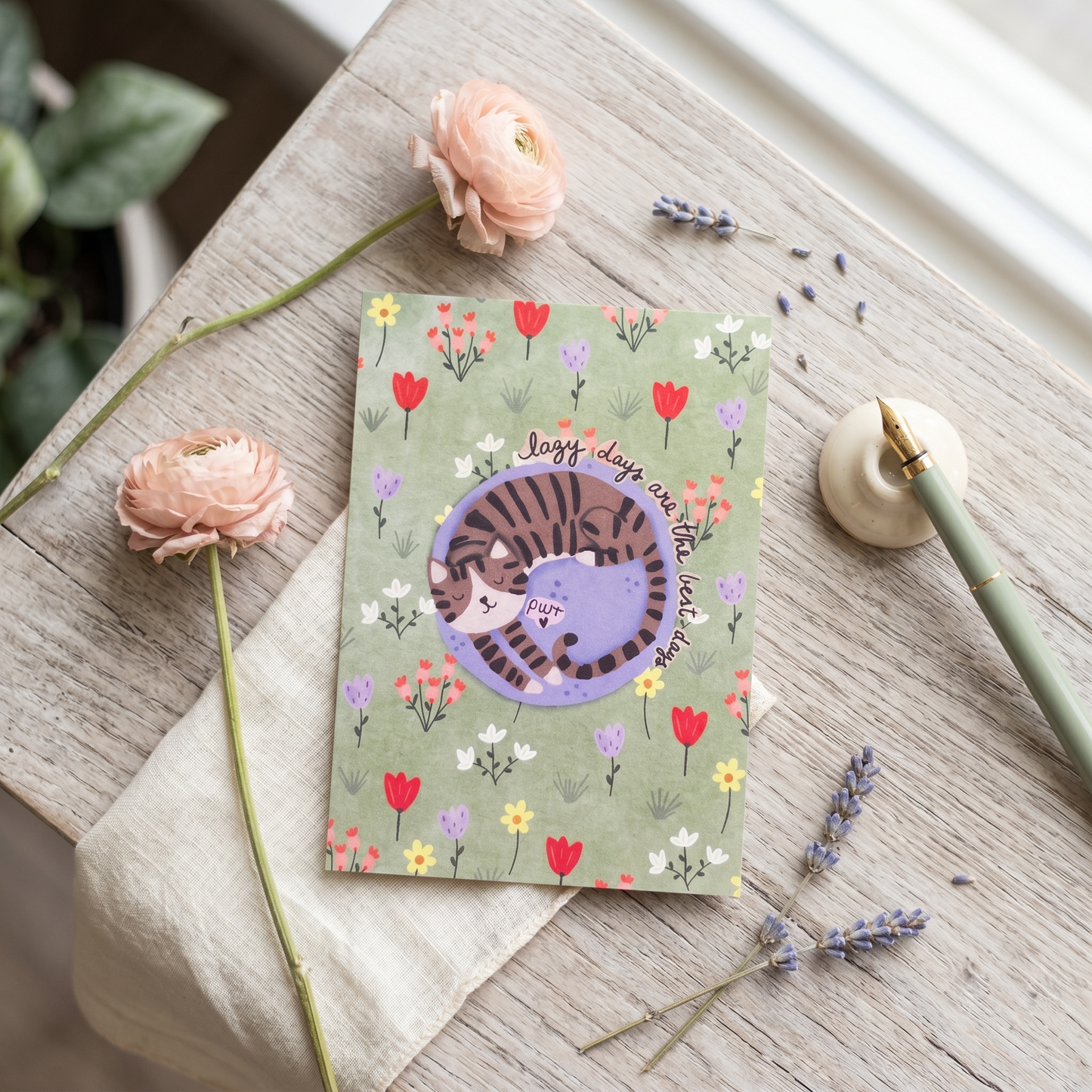 A square greeting card with a green floral background and a central illustration of a purple cat curled up under the words 'Jolly days come!' in cursive, styled on a rustic wooden table with pink flowers, lavender, and a green pen.