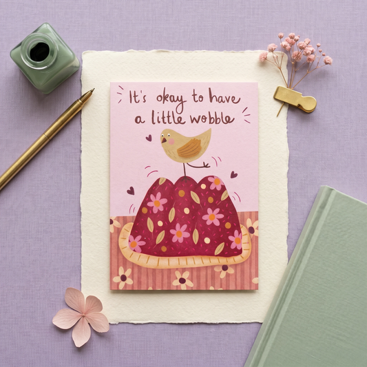 A square greeting card with a pink background and an illustration of a bird perched on a floral cake, reading 'It's okay to have a little wobble.' The card is styled in a flat lay on a lavender surface with a gold pen, green ink bottle, pink flowers, a clip, and a mint green notebook.