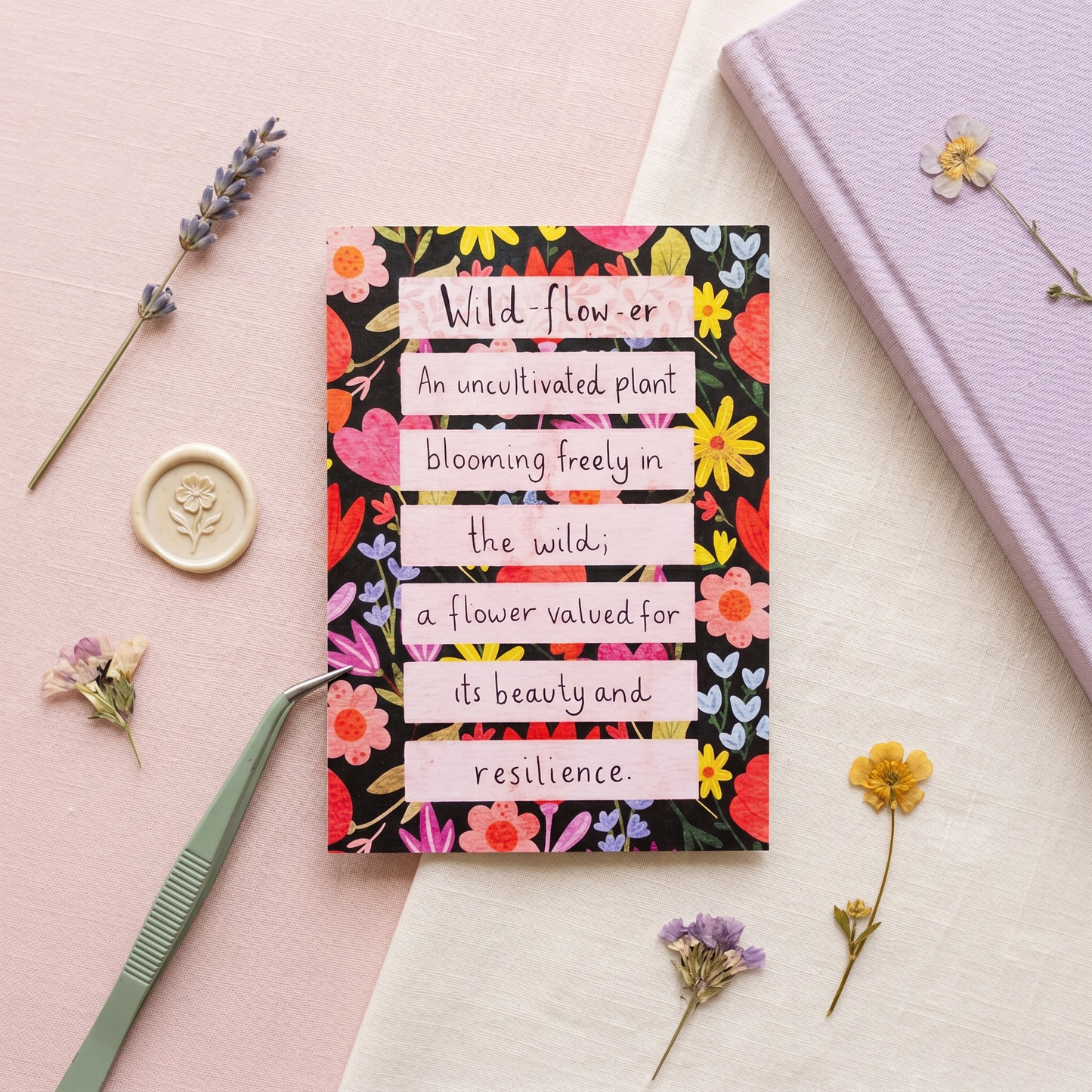 A square floral notebook or card with the quote 'Wild-flower: An uncultivated plant blooming freely in the wild; a flower valued for its beauty and resilience.' lies on a pink and white surface, styled with dried flowers, tweezers, a wax seal, and a lavender notebook.