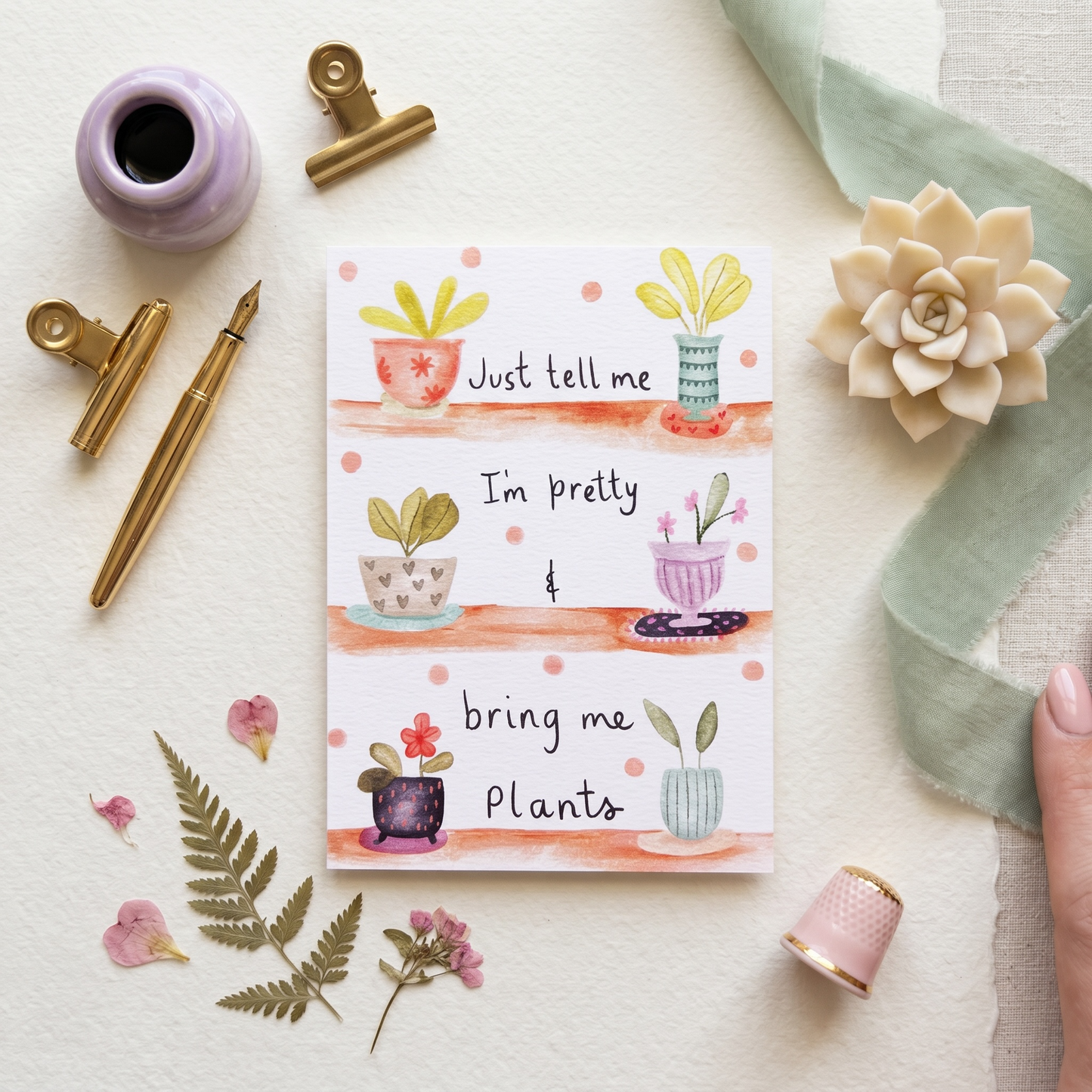 A flat-lay of a cute, illustrated stationery card with three rows of watercolor potted plants and the text 'Just tell me I'm pretty bring me Plants'. The card is styled on a textured cream surface with decorative props including a lavender ink bottle, gold clips and pen, a pink thimble, dried ferns, and a succulent.