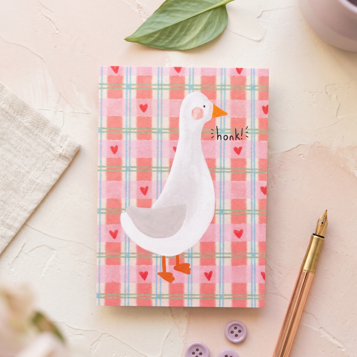 A square greeting card featuring a white goose on a pink plaid background with small red hearts and the word 'honk!' printed on it. The card is styled in a flat-lay arrangement on a textured cream surface with a green leaf, a beige cloth, a rose gold pen, and purple buttons.