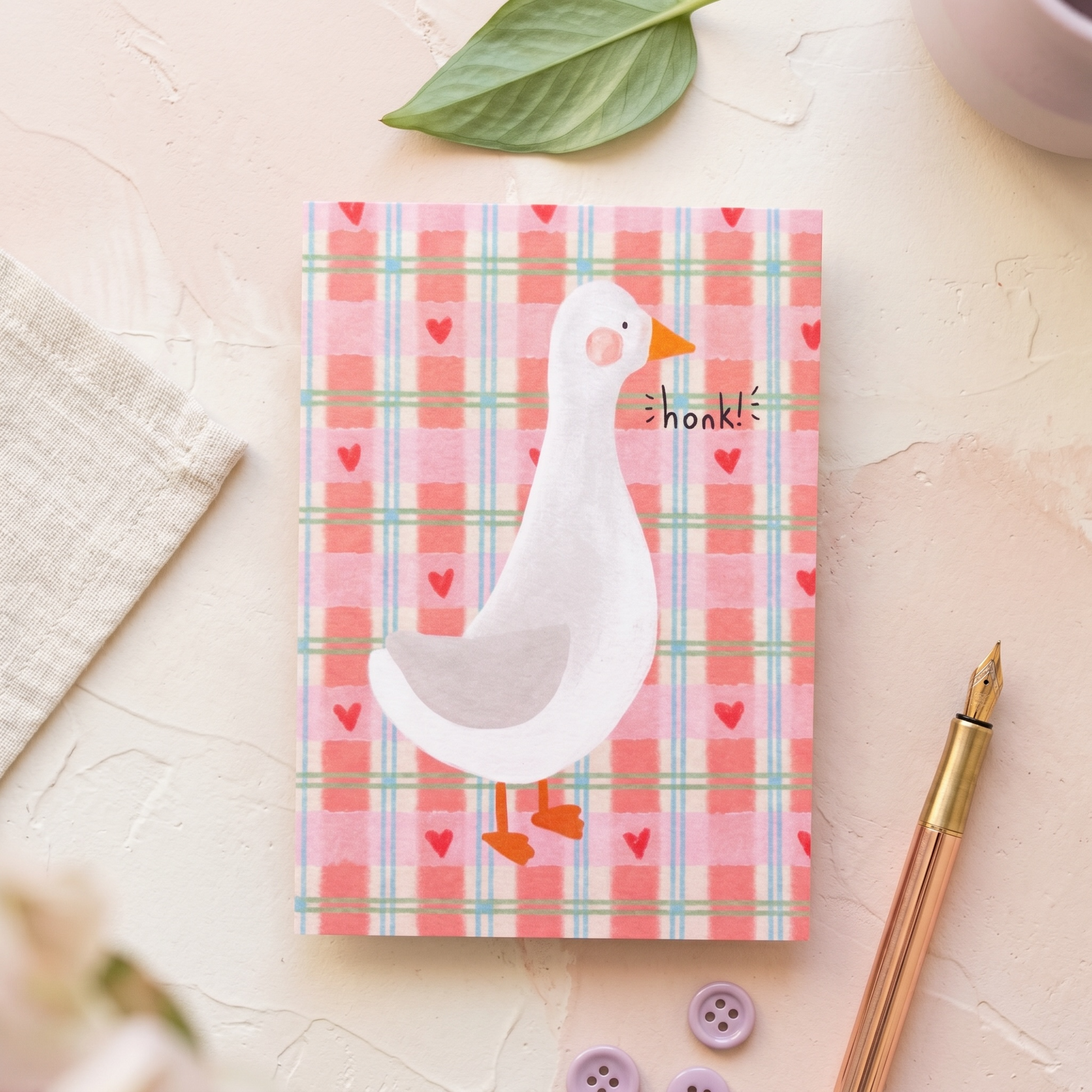 A square greeting card featuring a white goose on a pink plaid background with small red hearts and the word 'honk!' printed on it. The card is styled in a flat-lay arrangement on a textured cream surface with a green leaf, a beige cloth, a rose gold pen, and purple buttons.