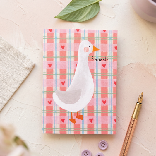 A square greeting card featuring a white goose on a pink plaid background with small red hearts and the word 'honk!' printed on it. The card is styled in a flat-lay arrangement on a textured cream surface with a green leaf, a beige cloth, a rose gold pen, and purple buttons.