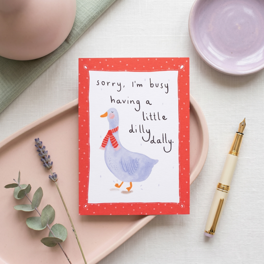 A greeting card with a red border and an illustration of a blue duck wearing a red striped scarf, placed on a pink tray with a pen, dish, and greenery around it. The card reads, 'sorry, i'm busy having a little dilly dally.