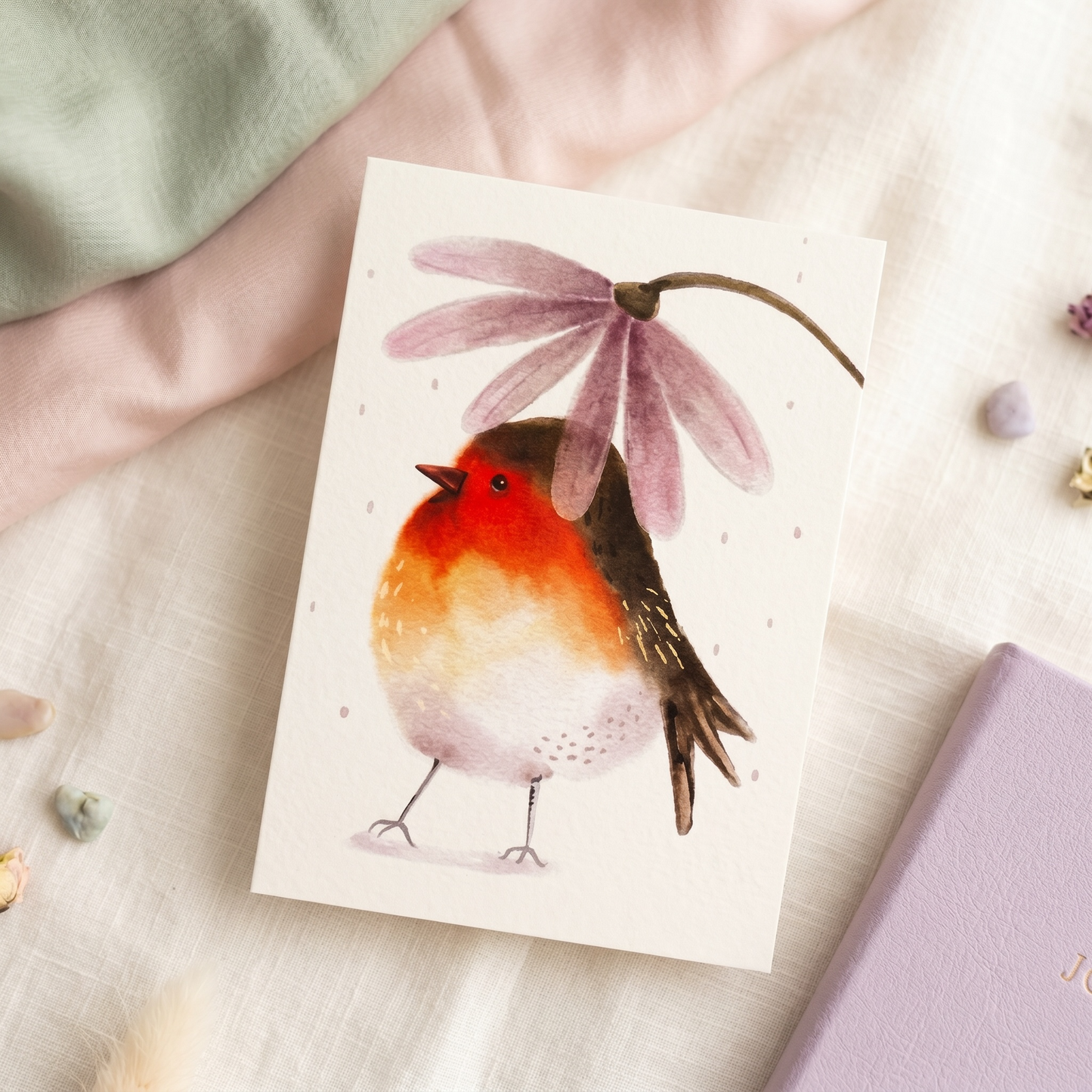 A square greeting card featuring a watercolor illustration of a robin bird under a pink flower, styled on a soft cream fabric with pastel props.