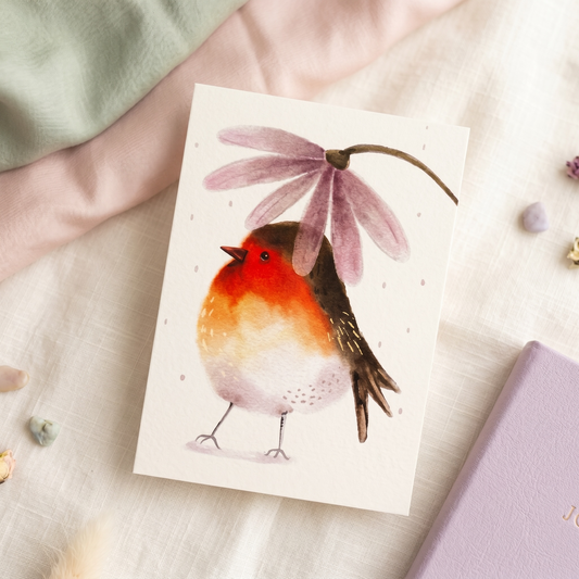 A square greeting card featuring a watercolor illustration of a robin bird under a pink flower, styled on a soft cream fabric with pastel props.