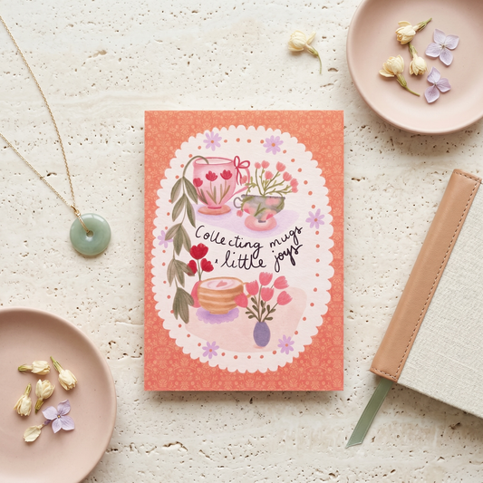 A coral pink notebook with a whimsical illustration of a teacup, flowers, and the text 'Collecting mugs, a little joy' is styled on a textured light stone surface with a gold necklace, a beige pen, and small dried flowers.