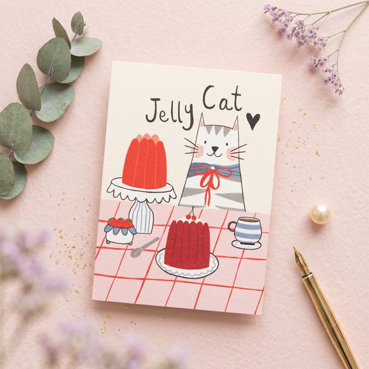 A cute illustrated greeting card titled 'Jelly Cat' lies on a pink surface with decorative eucalyptus leaves, small purple flowers, a pearl-like bead, and a gold pen. The card shows a gray-and-white cat wearing a red bow around its neck, standing beside red jelly cakes and a striped cup on a pink checkered tablecloth.