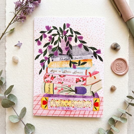 A watercolor-style art print featuring a stack of books with floral details, a purple flower bush, and decorative text such as 'Photo Album,' 'Home of Been Shades,' 'Journal,' 'Diary,' and 'Life Memories.' The print is photographed in a styled flat-lay on a textured cream surface with eucalyptus leaves, small stones, a wax seal, and a leather pen around it.