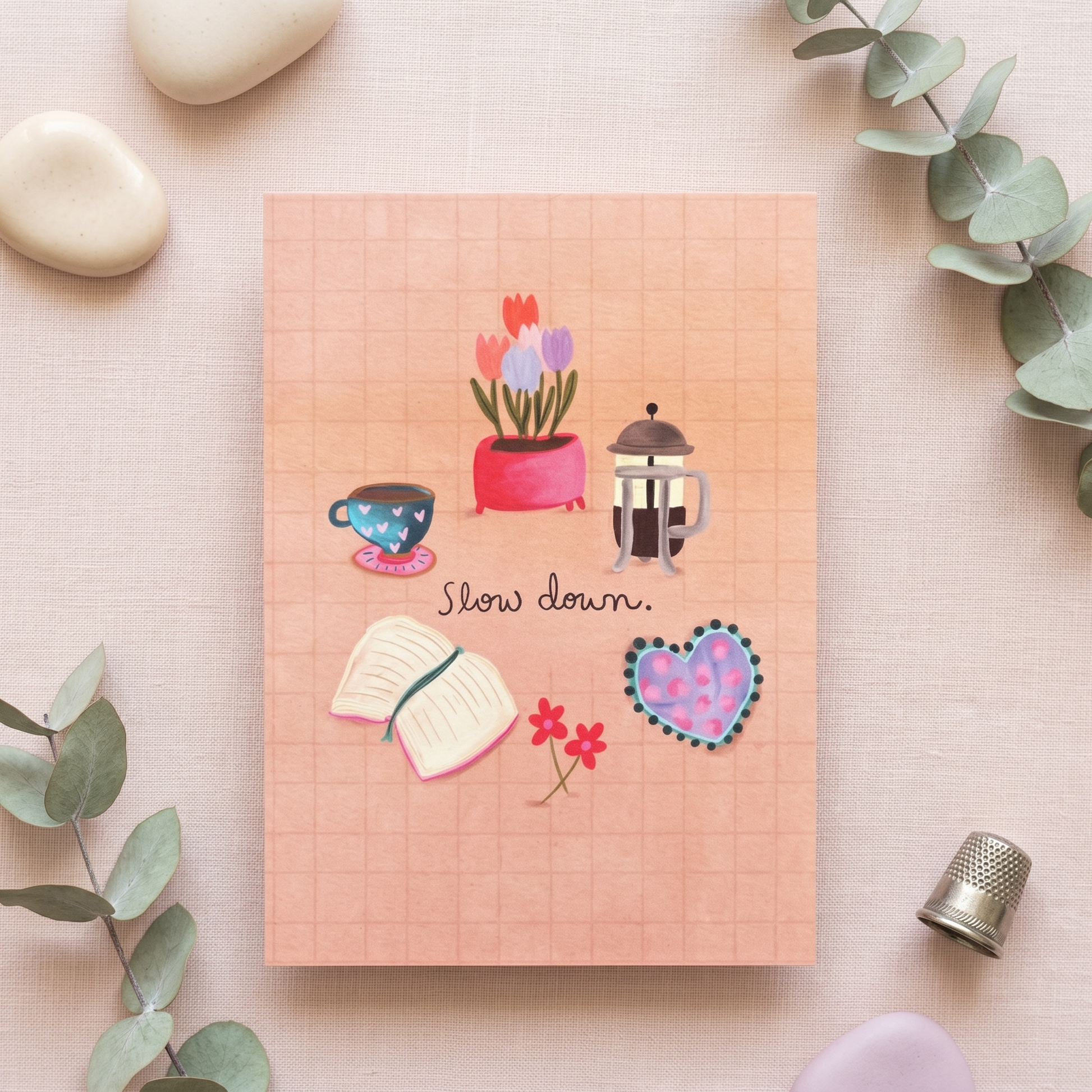 A square peach-colored notebook or card with a grid pattern, decorated with illustrations of a potted tulip, a teacup, a lantern, an open book, small red flowers, and a patterned heart. The center reads 'Slow down.