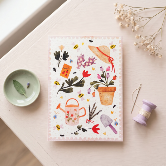 A square art print with a whimsical watercolor garden illustration, styled in a flat-lay on a light beige wooden table alongside a small dish, dried flowers, a lavender spool, and a needle.