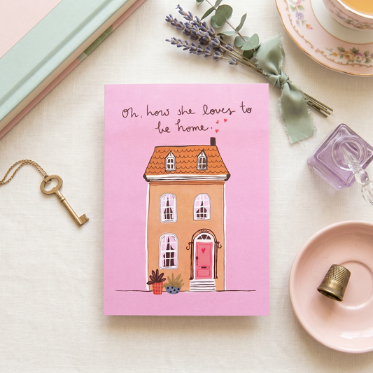 A pink greeting card featuring an illustration of a cozy house with the text 'Oh, how she loves to be home.' arranged in a styled flat-lay with a key, lavender, books, and a teacup.