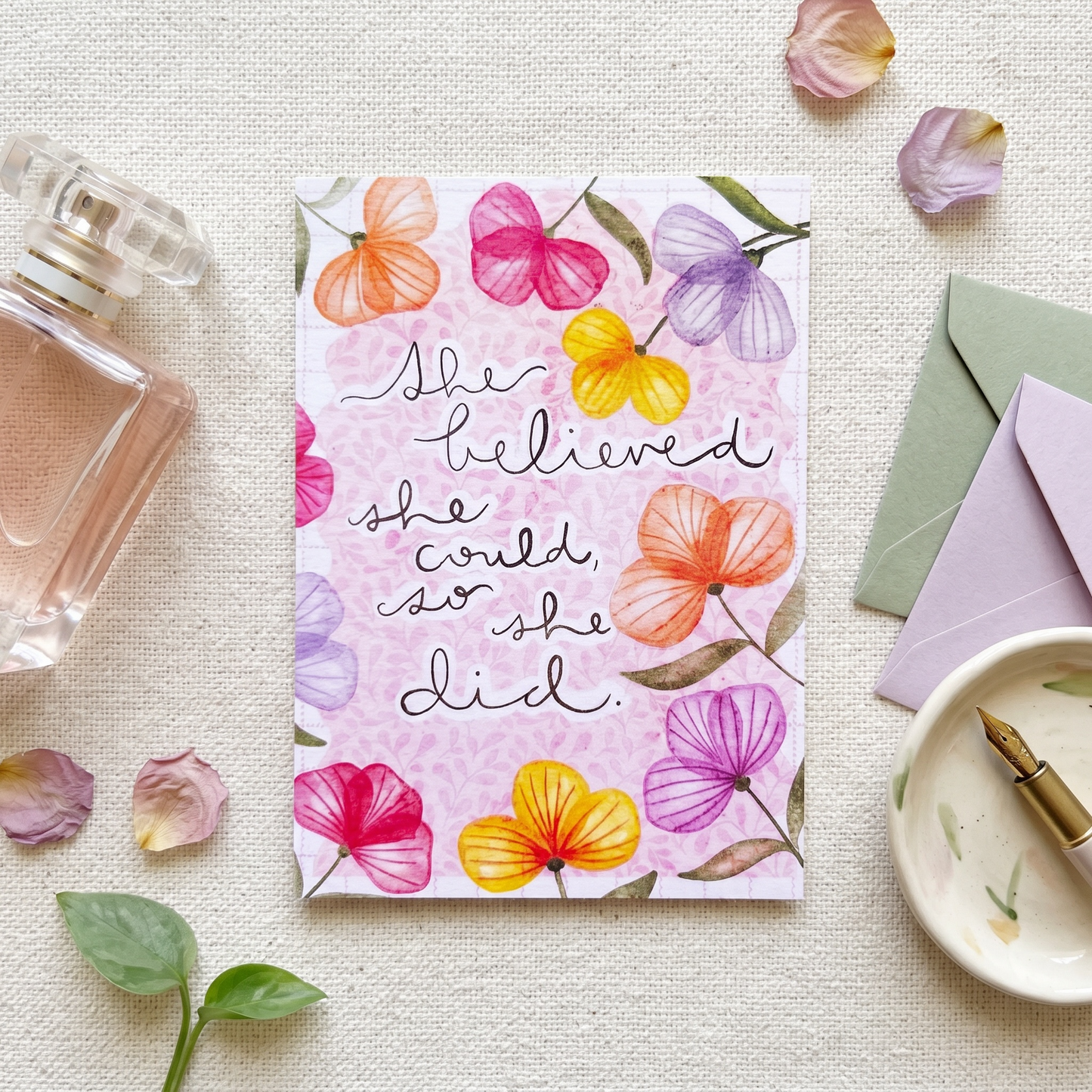 A floral art print card with the quote 'She believed she could, so she did.' in black script, styled in a flat-lay with a perfume bottle, envelopes, a dish, and scattered flower petals on a textured cream fabric background.