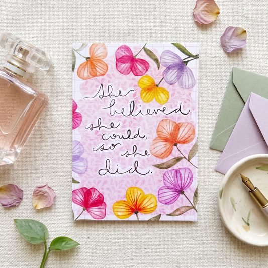 A floral art print card with the quote 'She believed she could, so she did.' in black script, styled in a flat-lay with a perfume bottle, envelopes, a dish, and scattered flower petals on a textured cream fabric background.