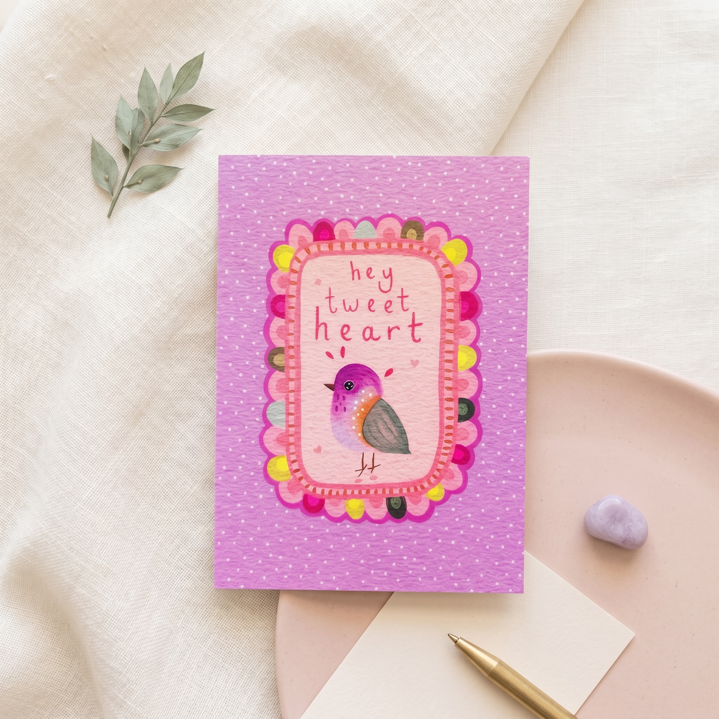 A pink greeting card with a decorative bird illustration and the text 'hey tweet heart' in a scalloped frame, styled on a neutral fabric surface with a small green sprig, a pink plate, a pen, and a lavender stone.