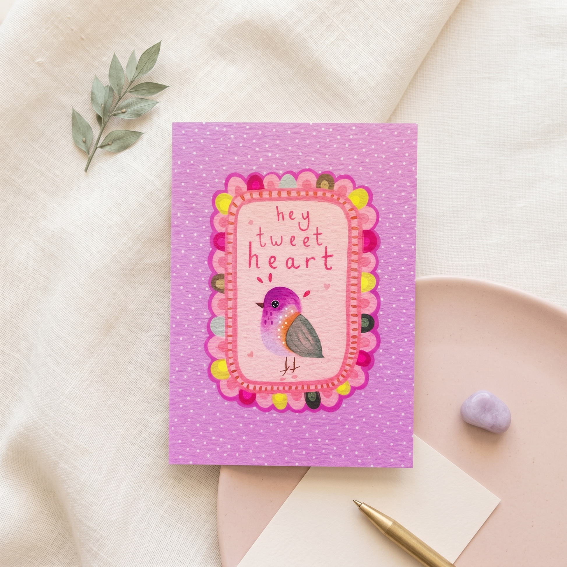 A pink greeting card with a decorative bird illustration and the text 'hey tweet heart' in a scalloped frame, styled on a neutral fabric surface with a small green sprig, a pink plate, a pen, and a lavender stone.