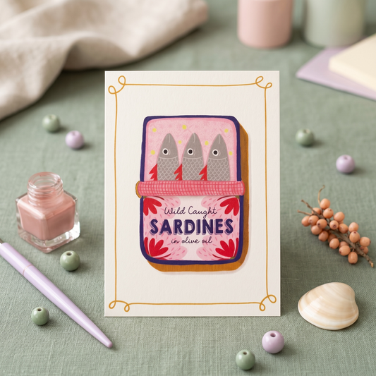 A square greeting card featuring an illustration of three gray sardines in a pink tin with the words 'Wild Caught Sardines in olive oil,' styled on a sage green surface with small beads, a pink bottle, a lavender pen, and soft neutral props in the background.