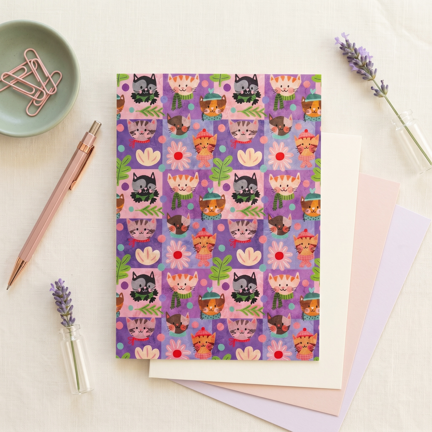 A flat-lay of a notebook with a colorful cartoon cat pattern on a purple cover, styled on a light fabric background with lavender sprigs, a rose gold pen, paper clips, and pastel sheets.