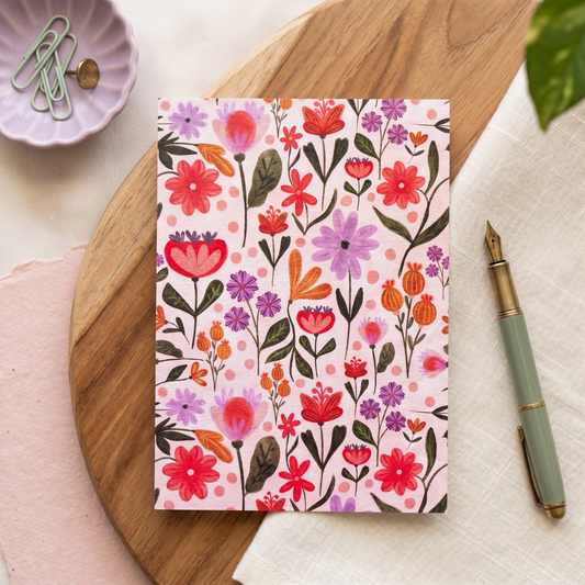 A floral-patterned notebook or card with a pink background and colorful watercolor flowers sits on a wooden board, styled with a green pen, paperclips, and a textured fabric surface.