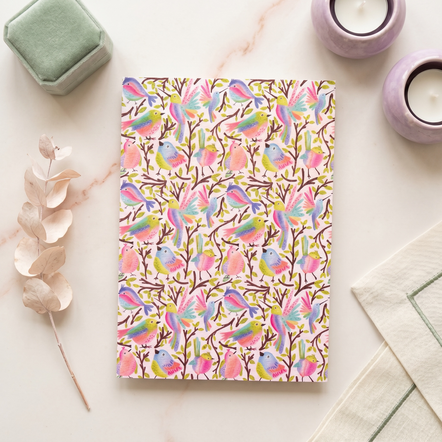 A notebook with a colorful watercolor bird pattern on a white cover, styled in a flat-lay arrangement with a green velvet box, lavender candles, dried eucalyptus, and a cream cloth on a light marble surface.