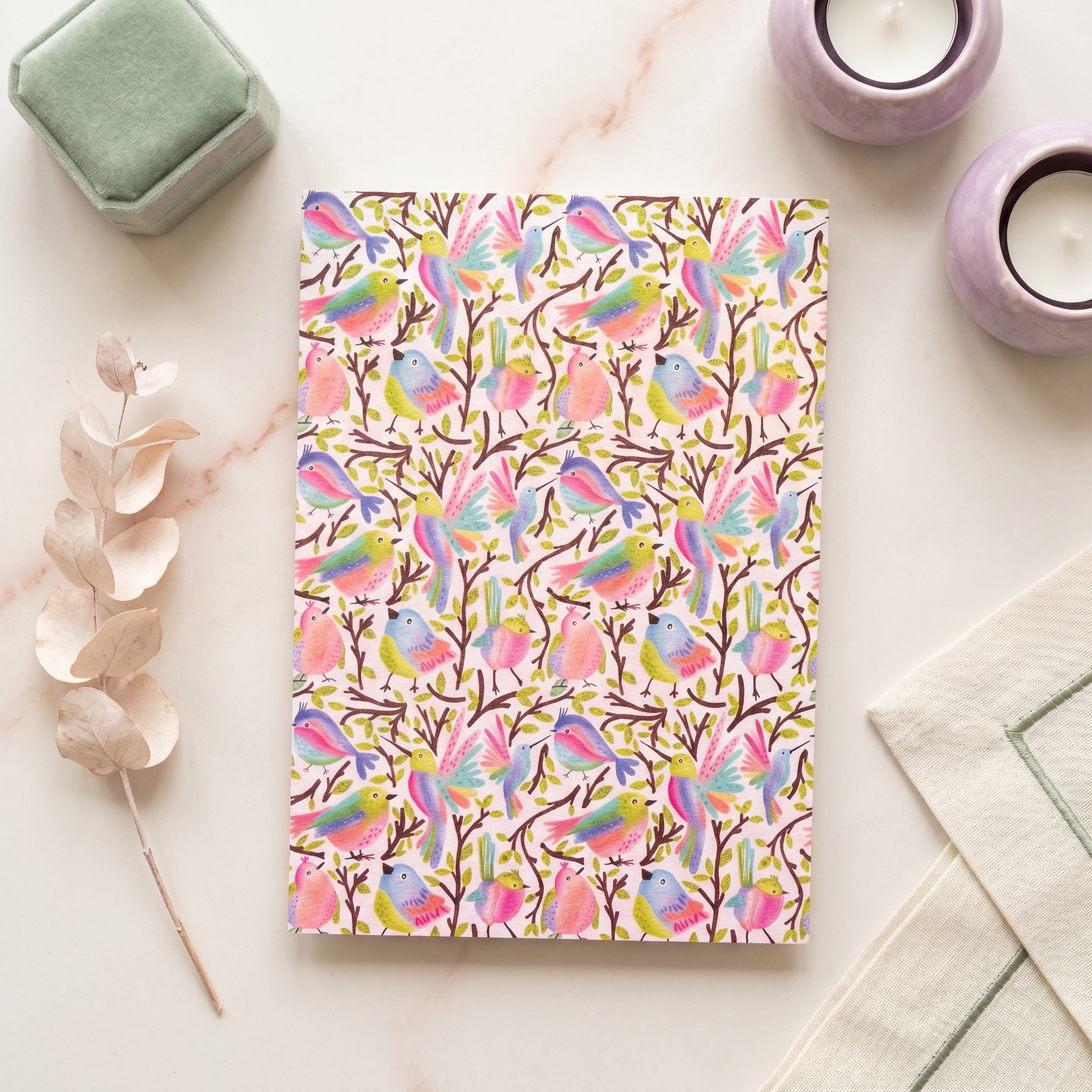 A notebook with a colorful watercolor bird pattern on a white cover, styled in a flat-lay arrangement with a green velvet box, lavender candles, dried eucalyptus, and a cream cloth on a light marble surface.