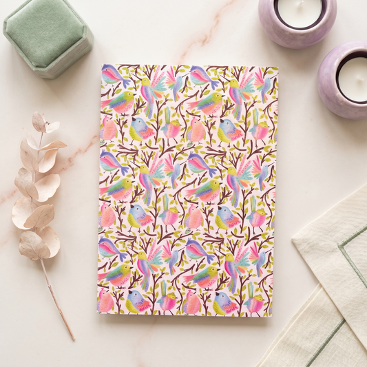 A notebook with a colorful watercolor bird pattern on a white cover, styled in a flat-lay arrangement with a green velvet box, lavender candles, dried eucalyptus, and a cream cloth on a light marble surface.