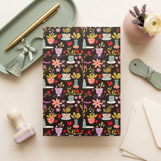 A flat-lay of a notebook with a dark charcoal background and a colorful pattern of potted plants, flowers, and leaves. The notebook is styled on a light beige surface with a mint green tray holding a gold pen and tassel, a pink vase with a white rose and lavender, a leather strap, a wooden stamp, and cream-colored envelopes.