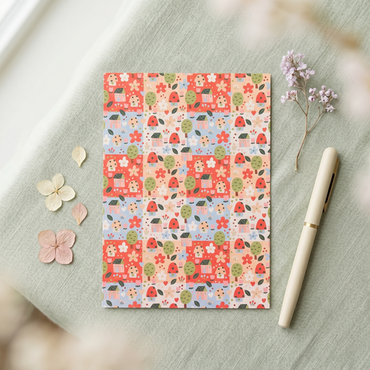 A flat sheet of patterned paper with a repeating patchwork design of red houses, green trees, and small floral motifs, styled on a pale green fabric surface with a cream pen and dried flowers nearby.